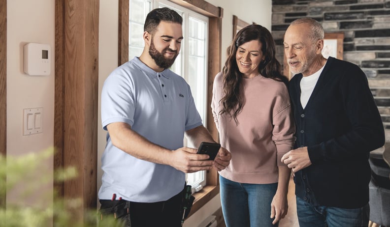 Un installateur Hilo avec des nouveaux clients Hilo regardent un téléphone dans une maison avec boiseries et grandes fenêtres; un thermostat intelligent est visible sur le mur.