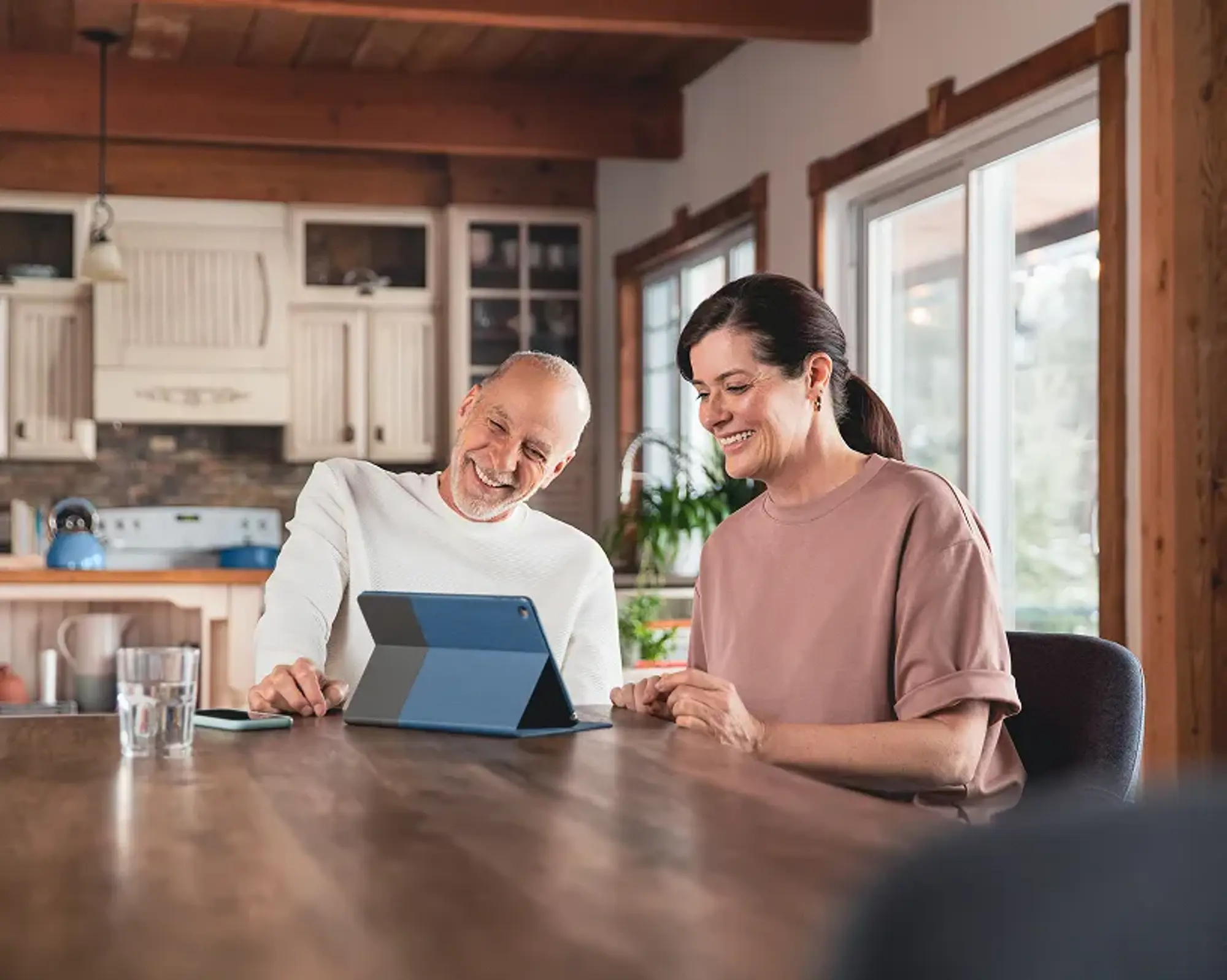 A couple sitting at a table, using a tablet with a blue case. A phone is placed next to it, with a Hilo thermostat behind them.