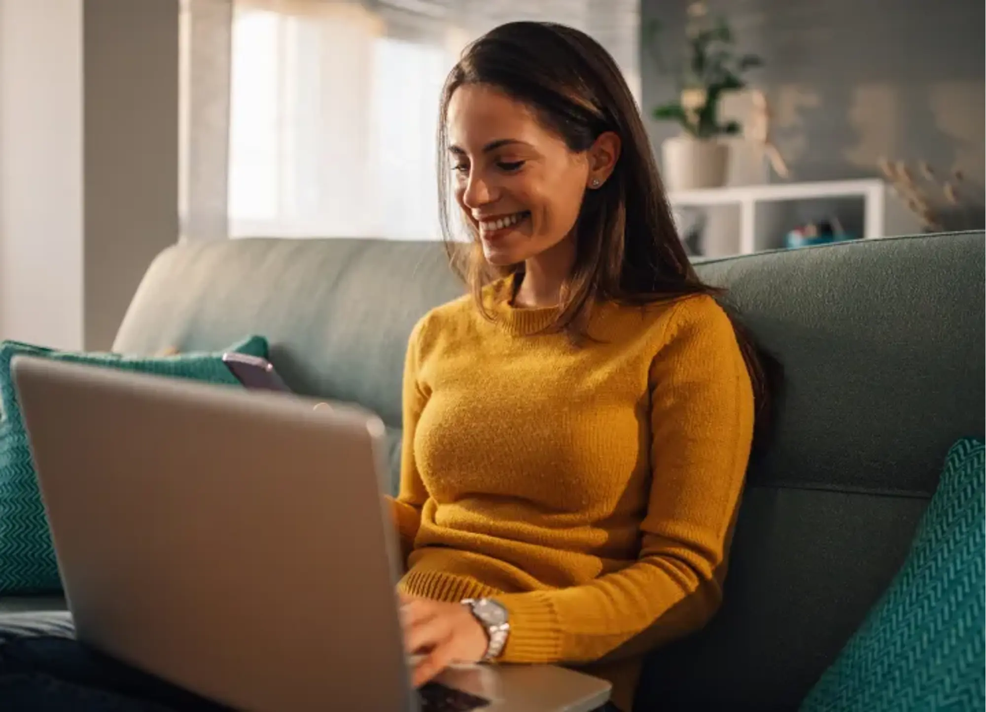 A smiling young woman sitting on a couch at home, using a laptop and holding a smartphone while speaking with Hilo customer support.