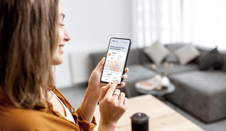 Person using the Hilo app on a smartphone to track their energy consumption, in a modern living room with a gray sofa and coffee table.
