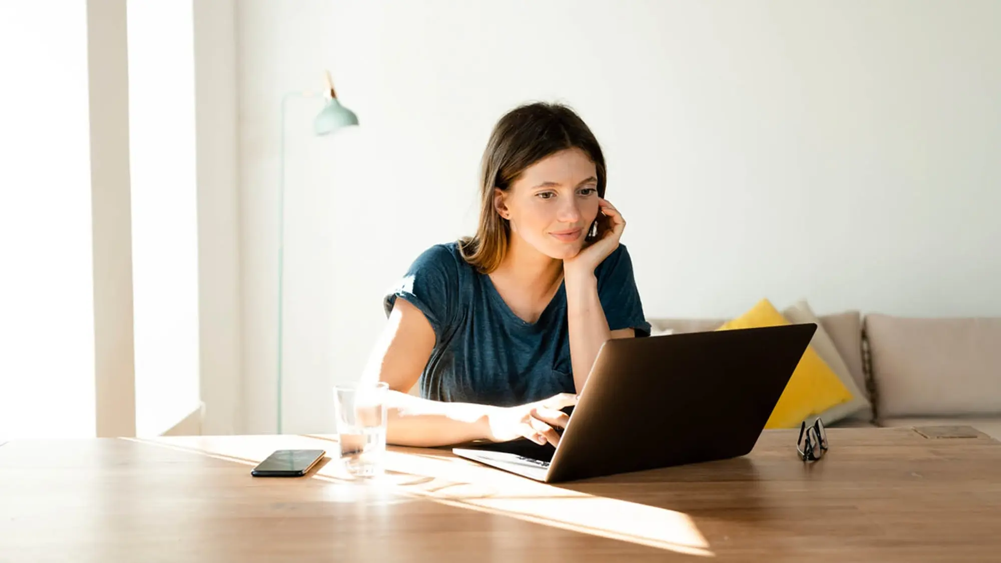 Femme assise à une table en bois, travaillant sur un ordinateur portable, avec un verre d’eau et un téléphone à côté, dans un intérieur lumineux.