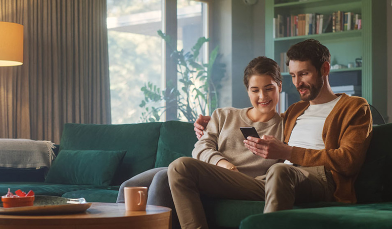 Un  couple souriant assis sur un canapé vert regardent un téléphone intelligent, dans un salon chaleureux avec lampe, étagères et table basse.