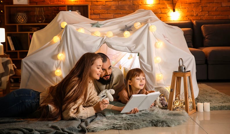 A family reads a book together inside a cozy blanket fort decorated with string lights and lanterns in a warmly lit living room.