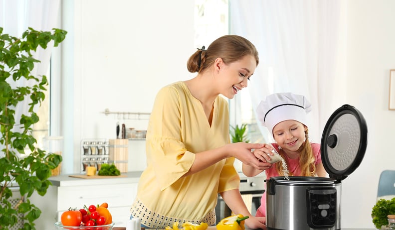 A mother and her daughter are cooking together; the child is wearing a chef's hat and adding ingredients to an electric cooker, with vegetables on the counter.