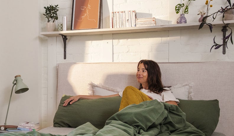 Une femme souriante assise dans un lit avec une couverture verte et un coussin jaune; mur en briques blanches avec étagère, livres et plantes.