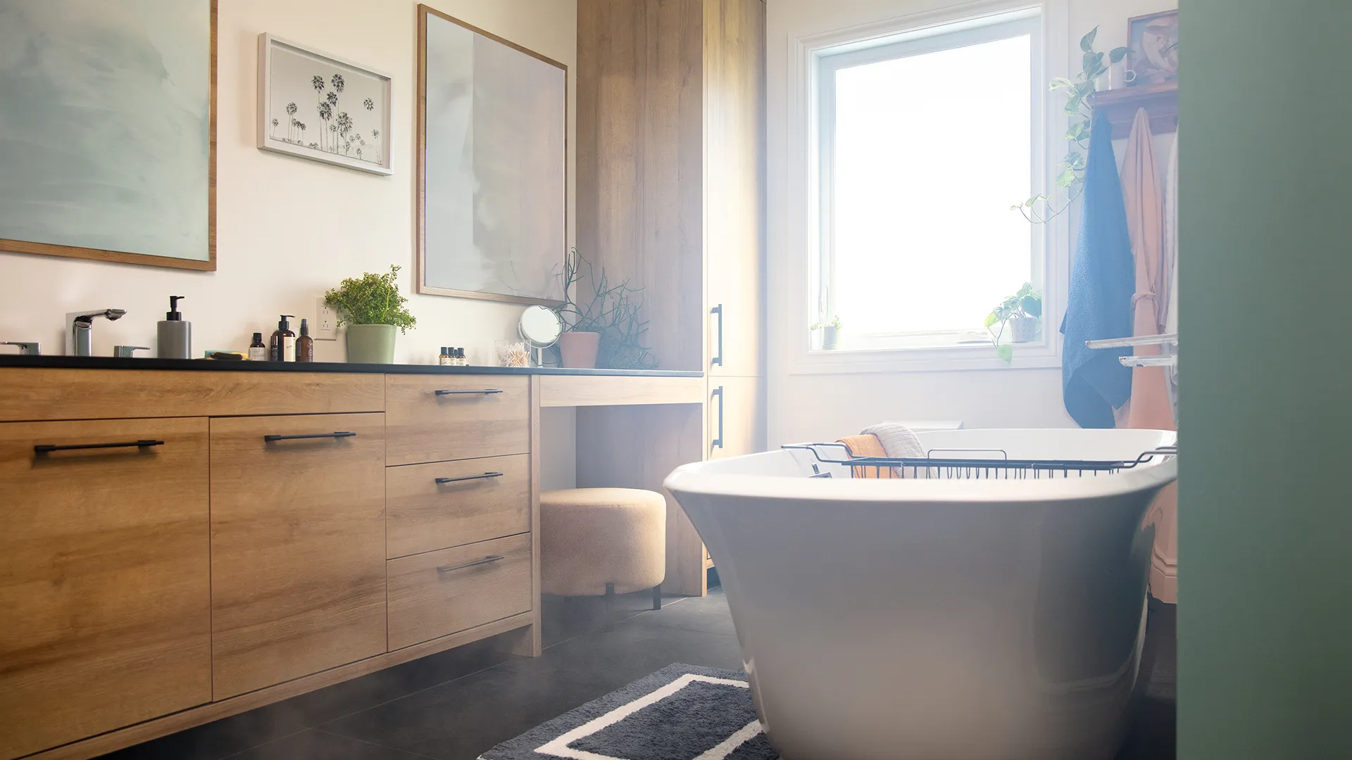 A bright bathroom with a white freestanding bathtub, a wooden vanity, decorative plants, and a large window that lets in natural light.