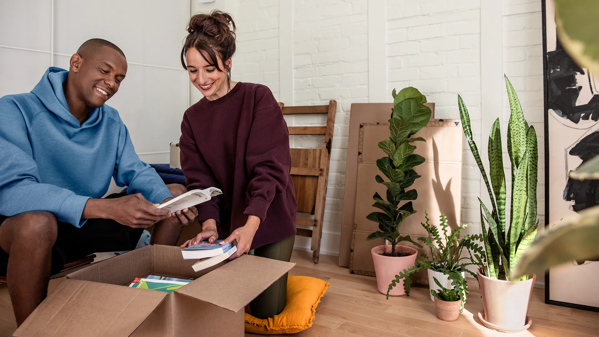 Deux nouveaux clients Hilo trient des livres dans une boîte de déménagement dans un salon lumineux, entourées de plantes d’intérieur et de cartons.