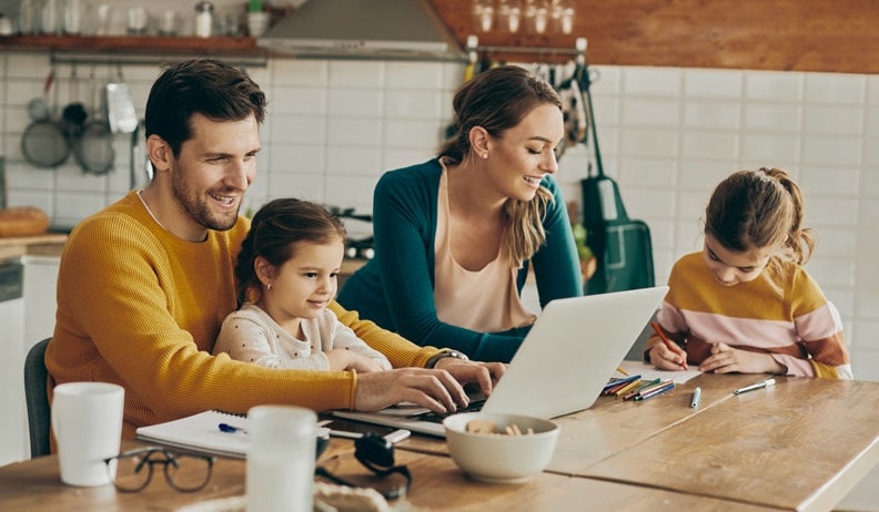 Family gathered around a counter: a man is helping a girl with a laptop, a woman is watching, another girl is drawing with colored pencils.