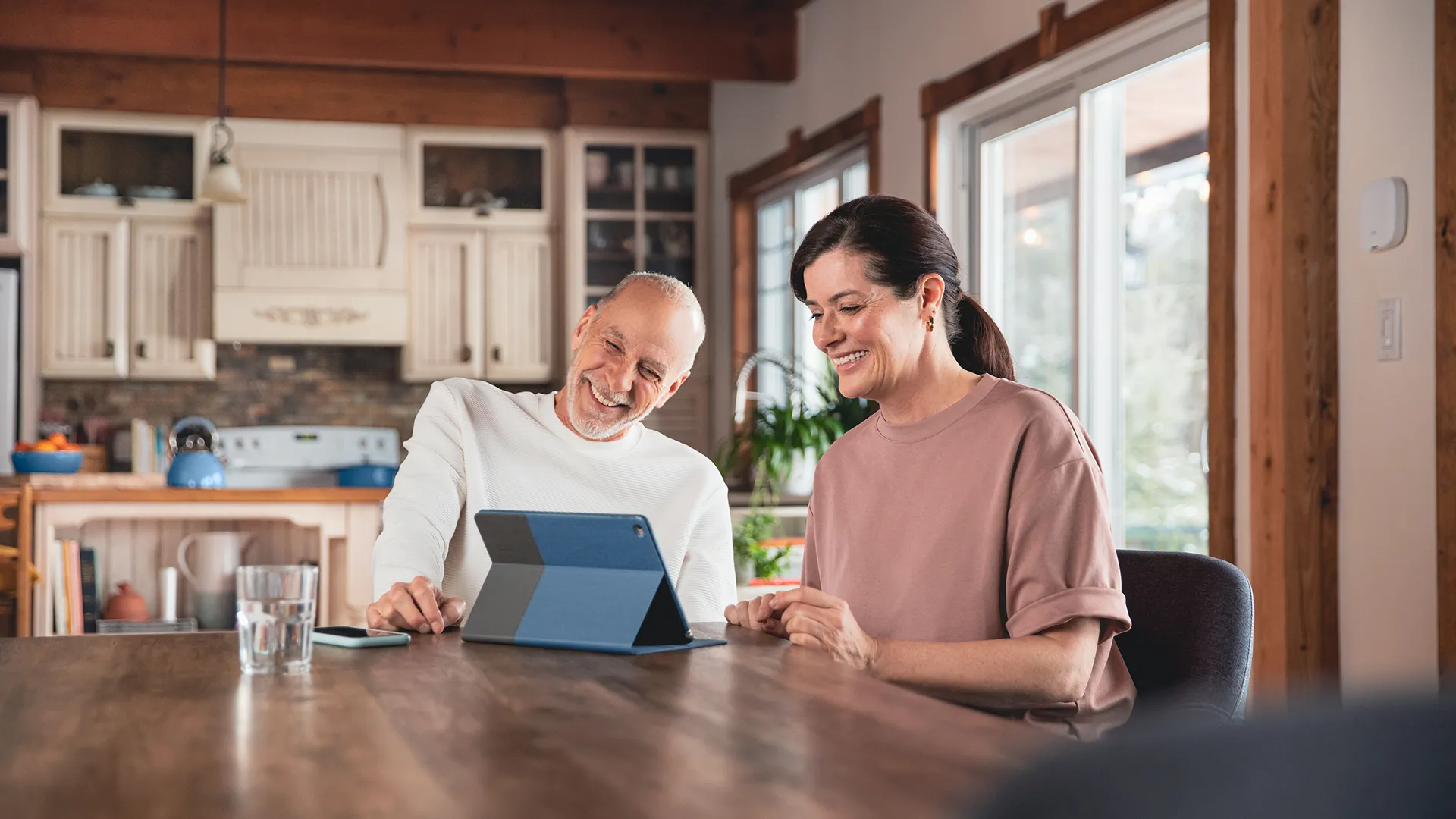A couple sitting at a table, using a tablet with a blue case. A phone is placed next to it, with a Hilo thermostat behind them.