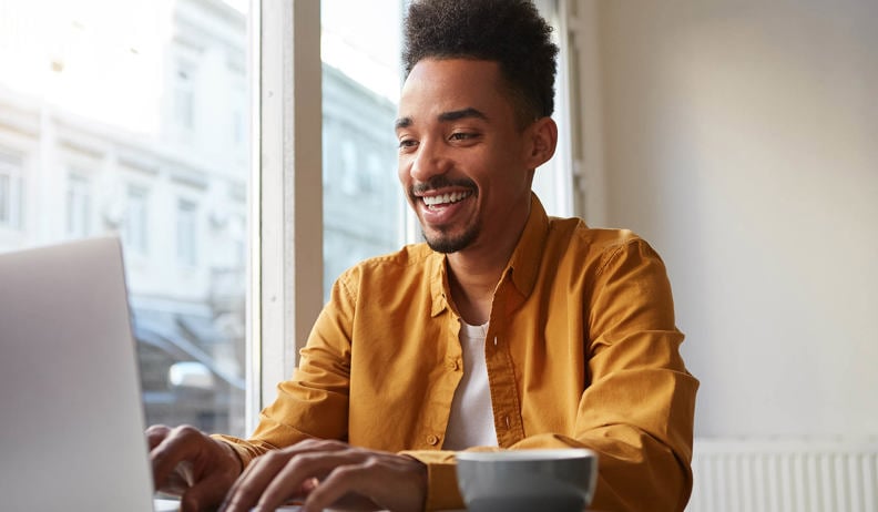Un homme souriant assis à une table utilise un ordinateur portable, porte une chemise moutarde; tasse à côté, lumière naturelle par la fenêtre.