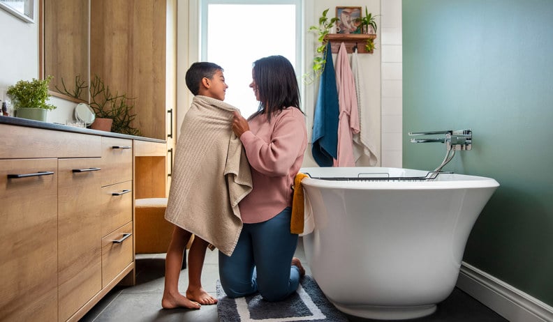 Une mère souriante est agenouillé dans une salle de bain, enveloppant un enfant dans une serviette beige.