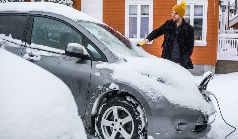 Un homme avec une tuque orange déneige une voiture électrique grise branchée devant une maison orange, sous la neige.