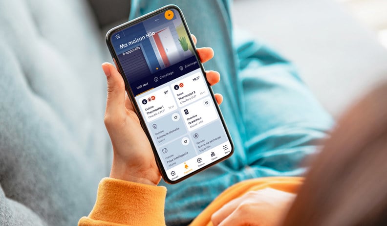 Personne tenant un téléphone affichant l'app Hilo avec des contrôles pour lumières, thermostats et autres appareils connectés.