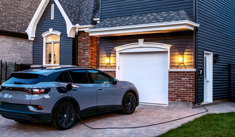 VUS électrique en charge dans une entrée privée, à côté d’une maison moderne avec revêtement foncé et porte de garage blanche.