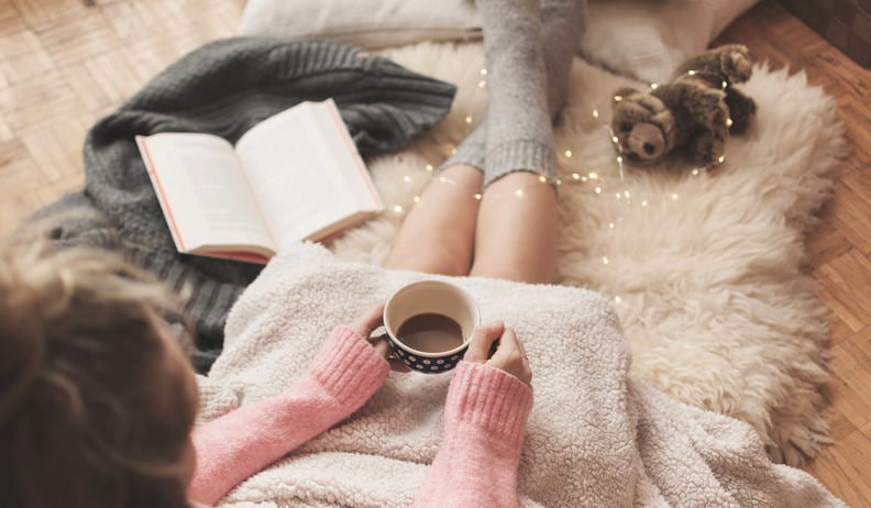 Une personne est assise sur un tapis moelleux avec une couverture, tenant une tasse de café. Elle porte un pull rose et des chaussettes confortables. Un livre ouvert et un ours en peluche sont à côté, avec des guirlandes lumineuses.