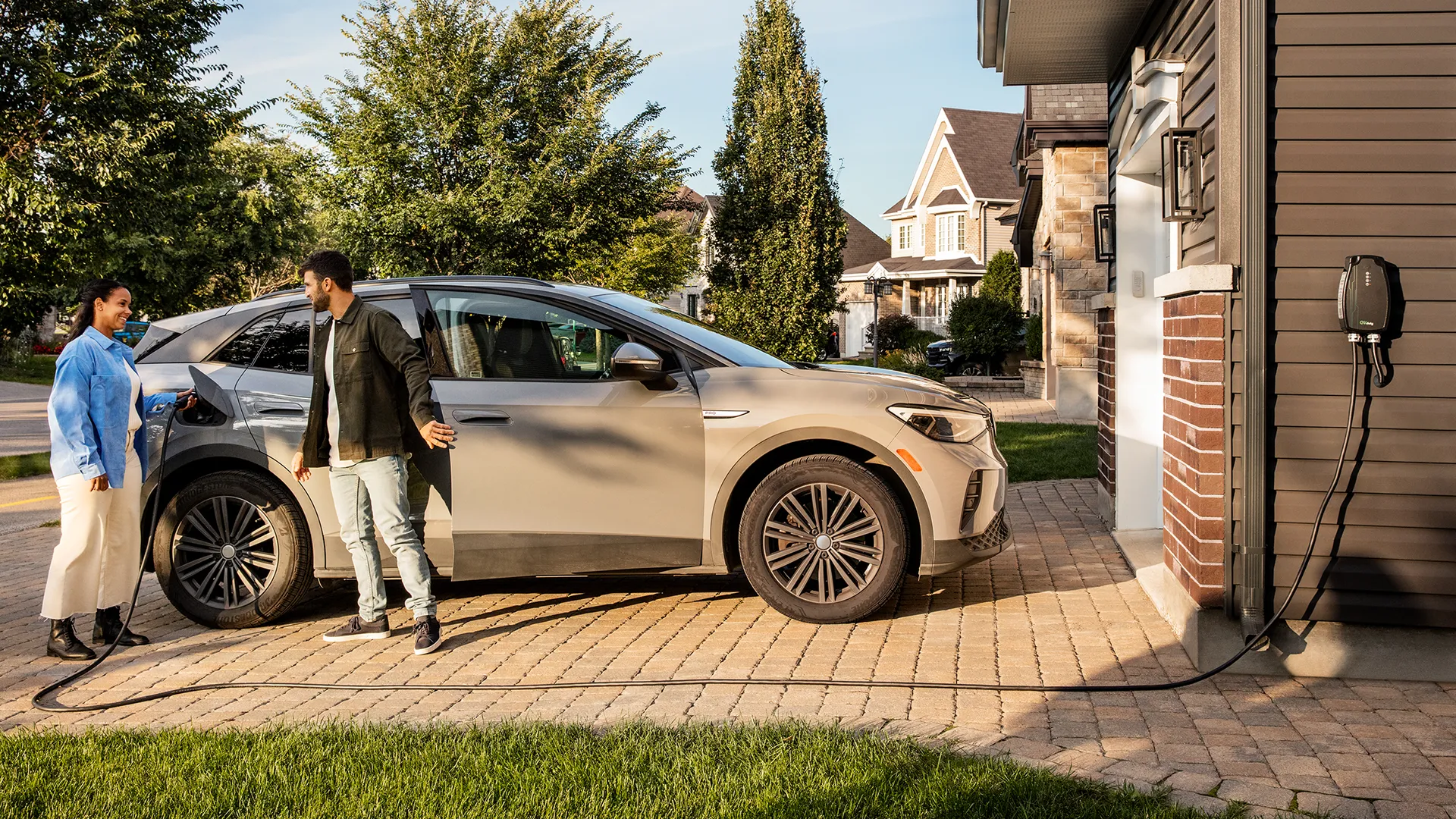 An electric vehicle plugged into a residential charging station in a driveway. Two people are standing near the vehicle.