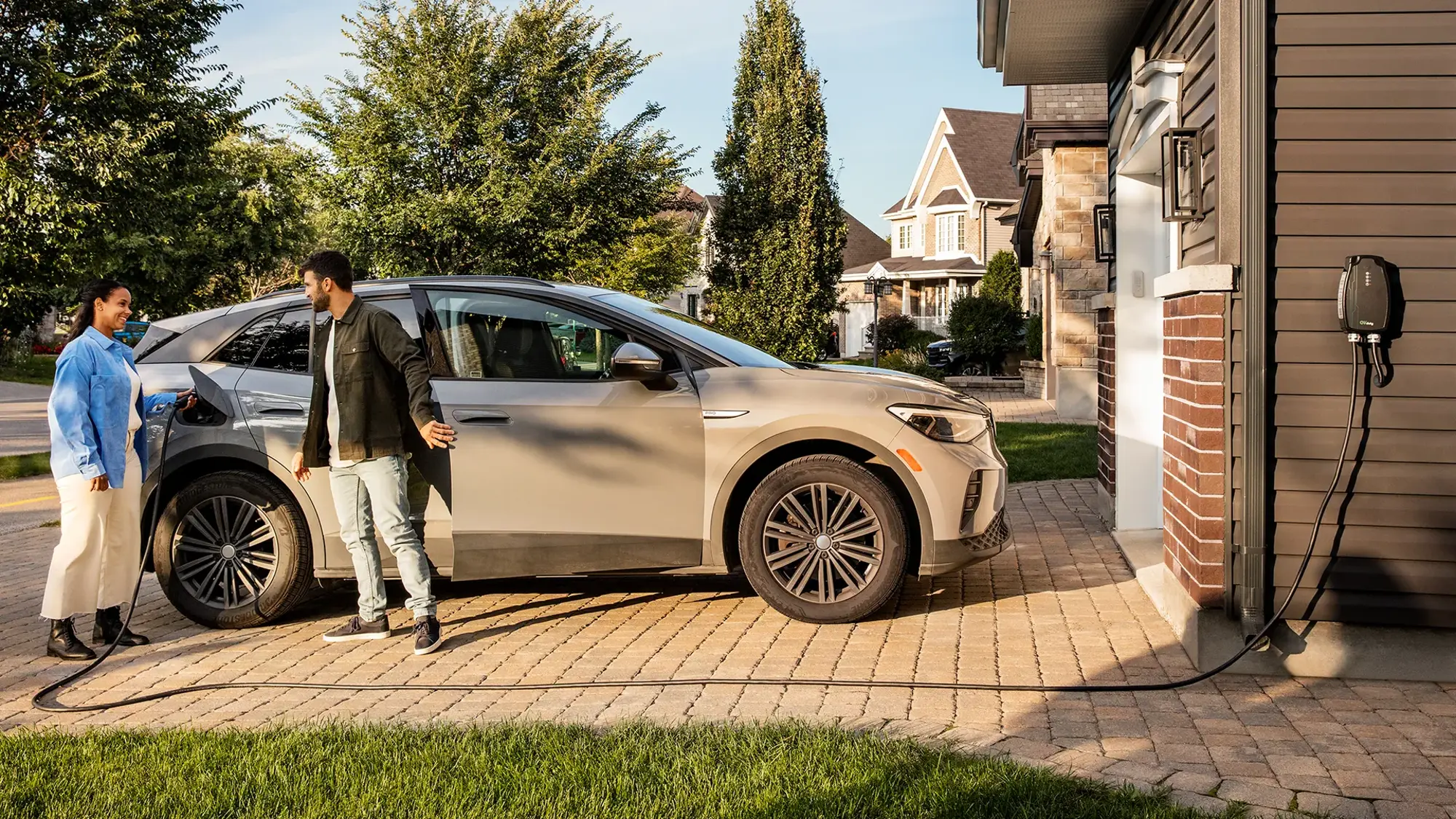 An electric vehicle plugged into a residential charging station in a driveway. Two people are standing near the vehicle.