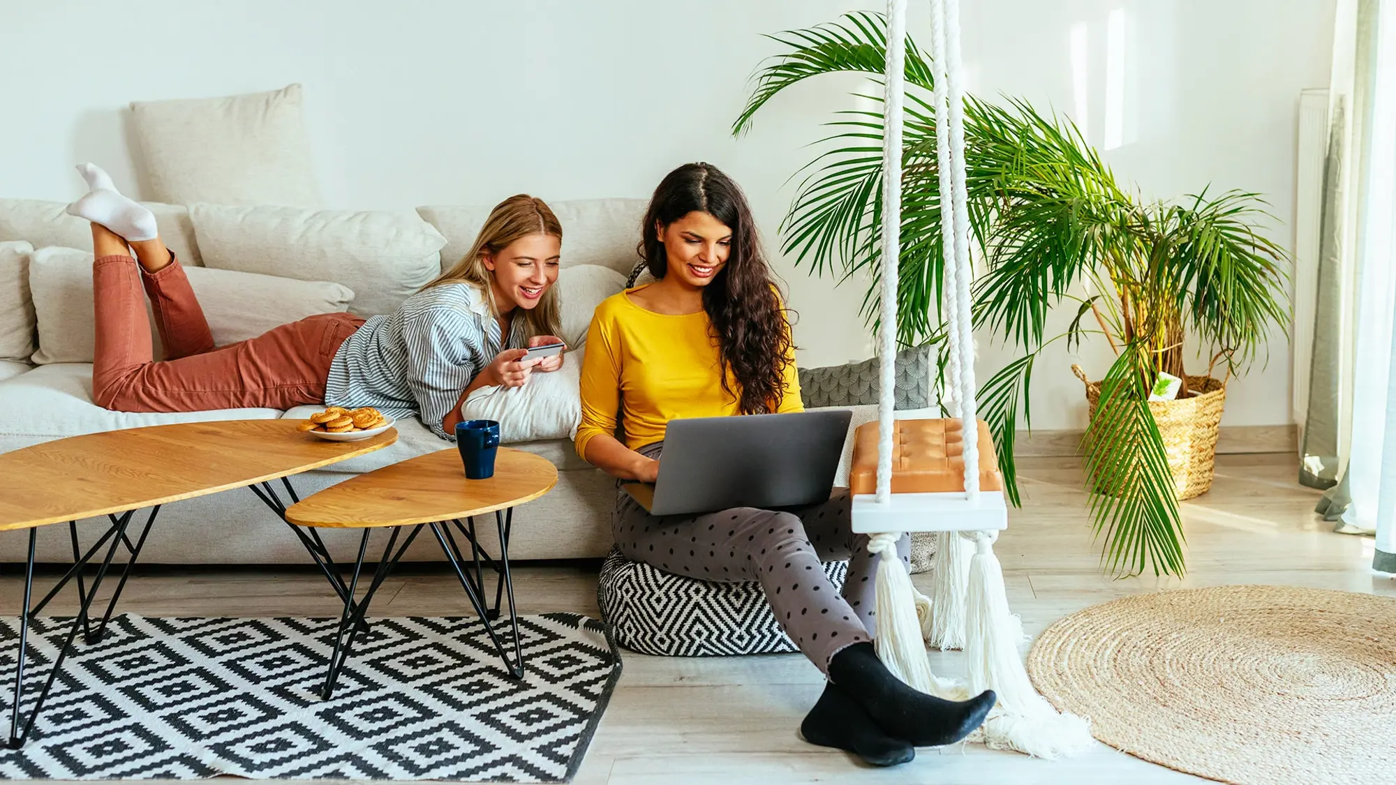 Two smiling people in a modern living room: one on a couch using a smartphone, the other seated on the floor with a laptop.