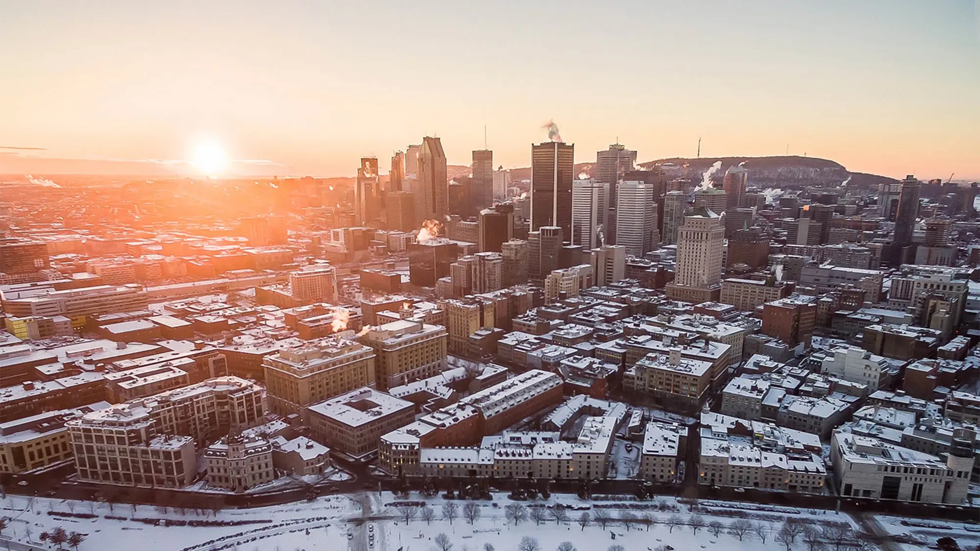 Aerial view of a snowy Montréal at sunrise, with tall skyscrapers, snow-covered streets, and a distant mountain in the background.