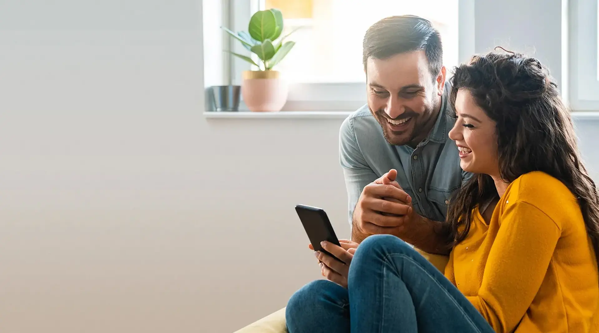 A smiling couple sitting on a sofa, looking at a smartphone together.