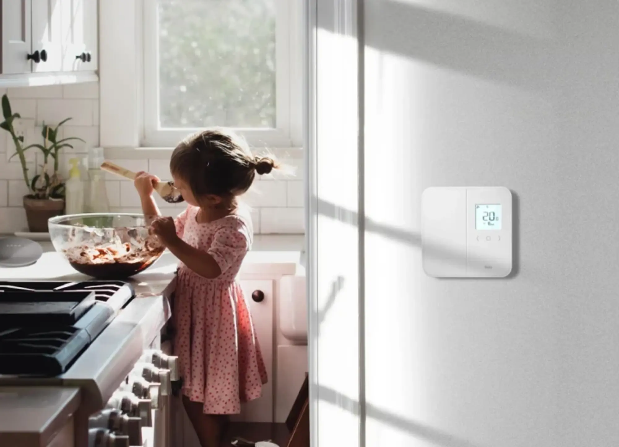 Jeune enfant mélangeant des ingrédients dans un bol sur le comptoir d’une cuisine ensoleillée, avec un thermostat intelligent affichant 20 degrés installé sur le mur.