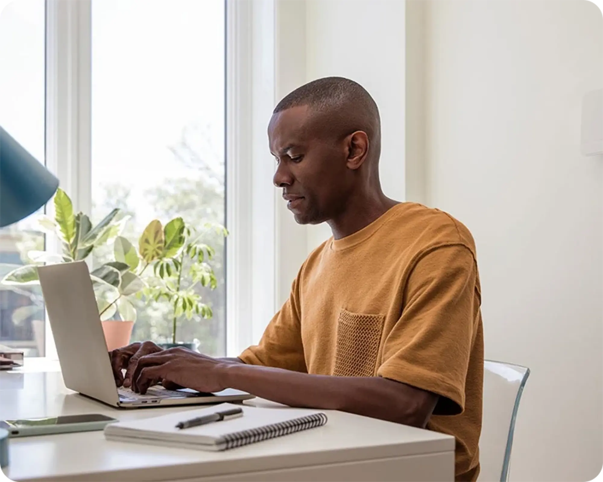 A man sitting at a white desk, typing on a laptop, in front of a bright window and a green plant.