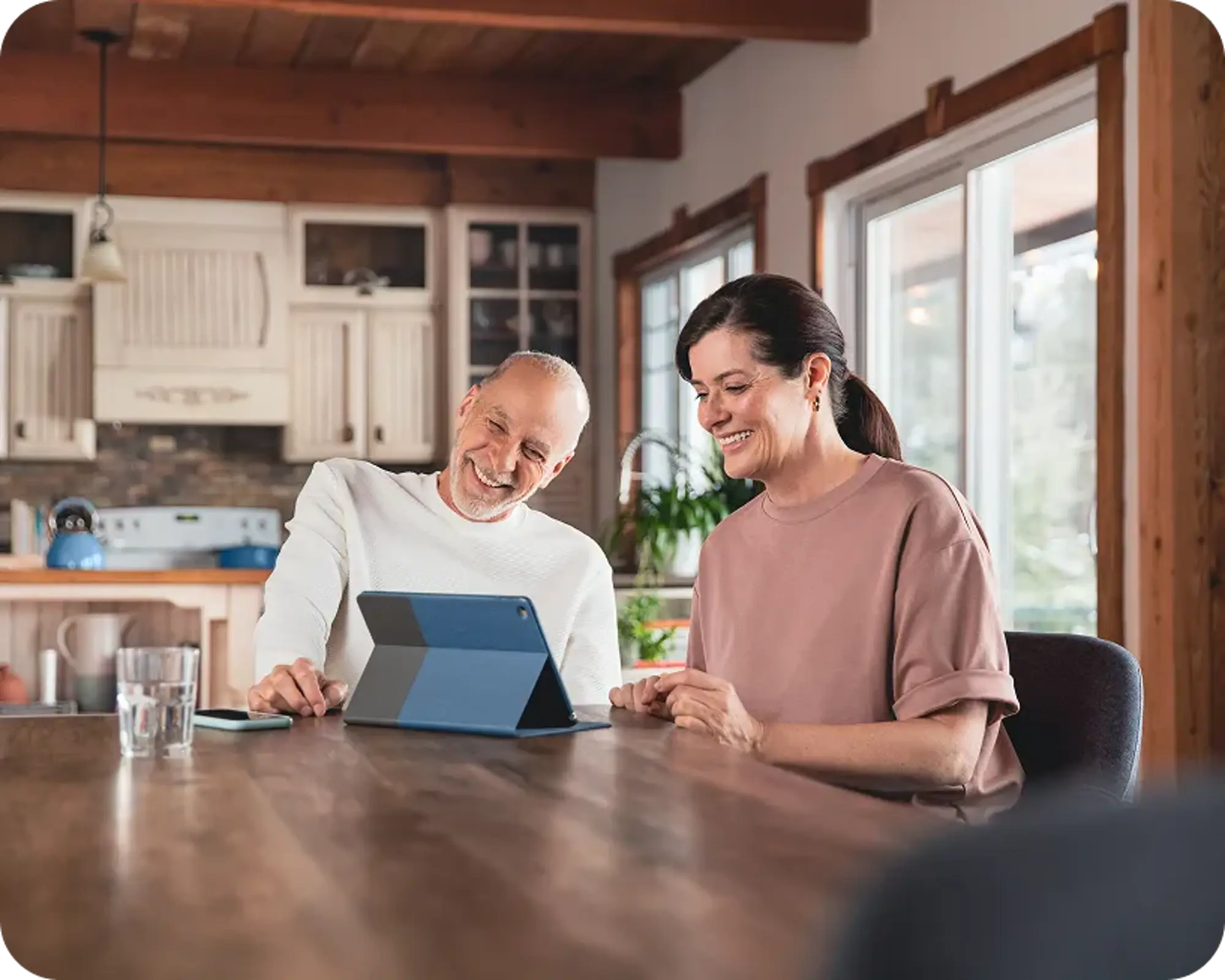 Un couple assis à une table, utilisant une tablette avec un étui bleu. Un téléphone est posé à côté, avec un thermostat Hilo derrière eux.